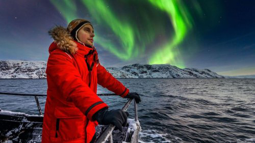 Man wearing red parka looks out at sea from the railing of a boat in front of a green aurora borealis