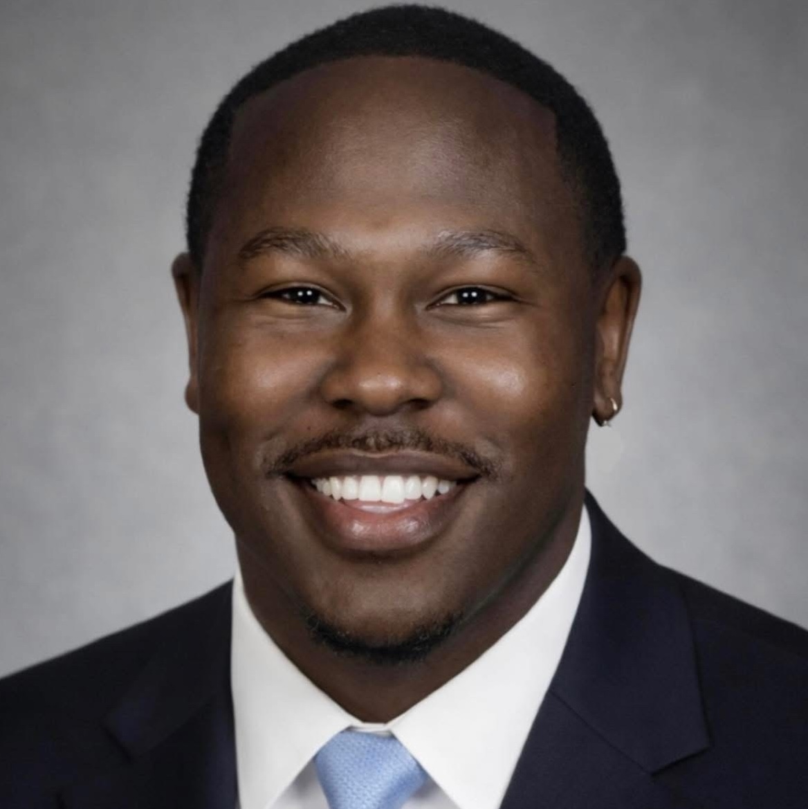Professional head-and-shoulders portrait of a smiling man in a dark suit and light blue tie against a gray backdrop.