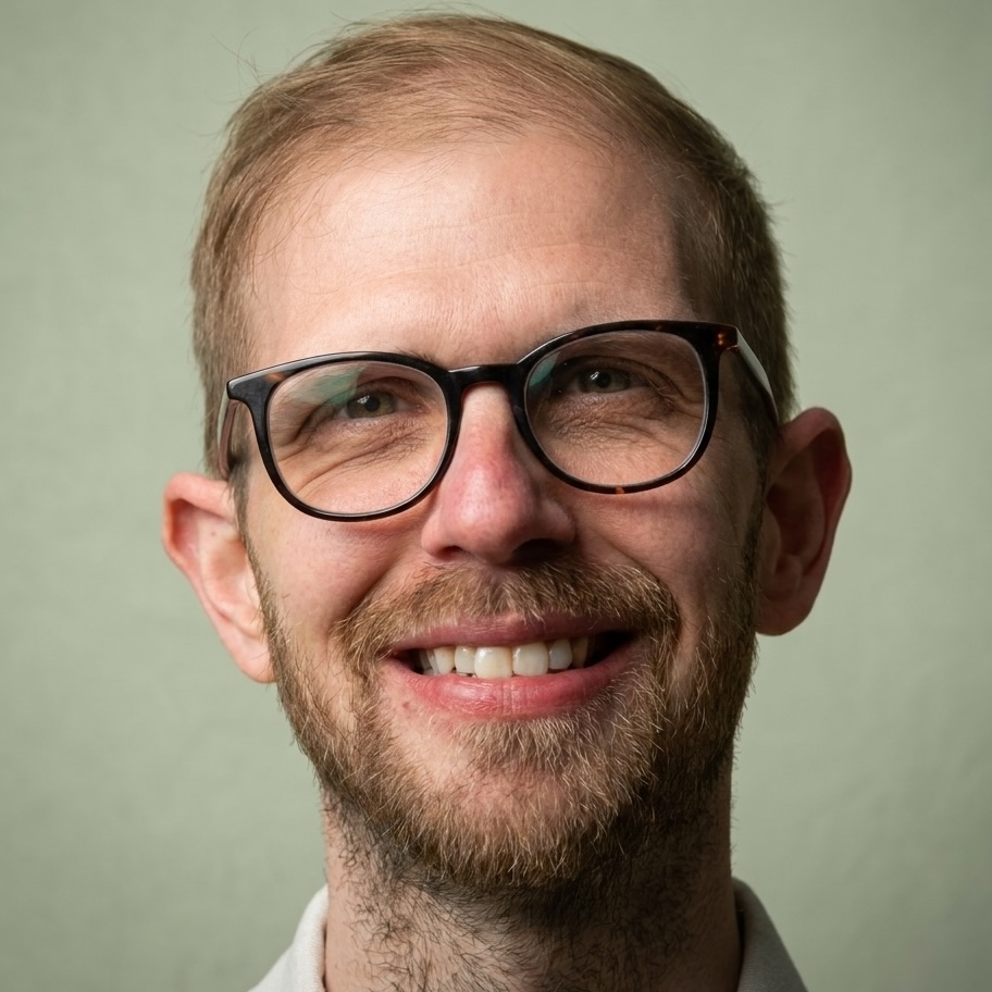 Smiling man with light brown hair and a short beard wearing round glasses, looking at the camera against a pale green background.