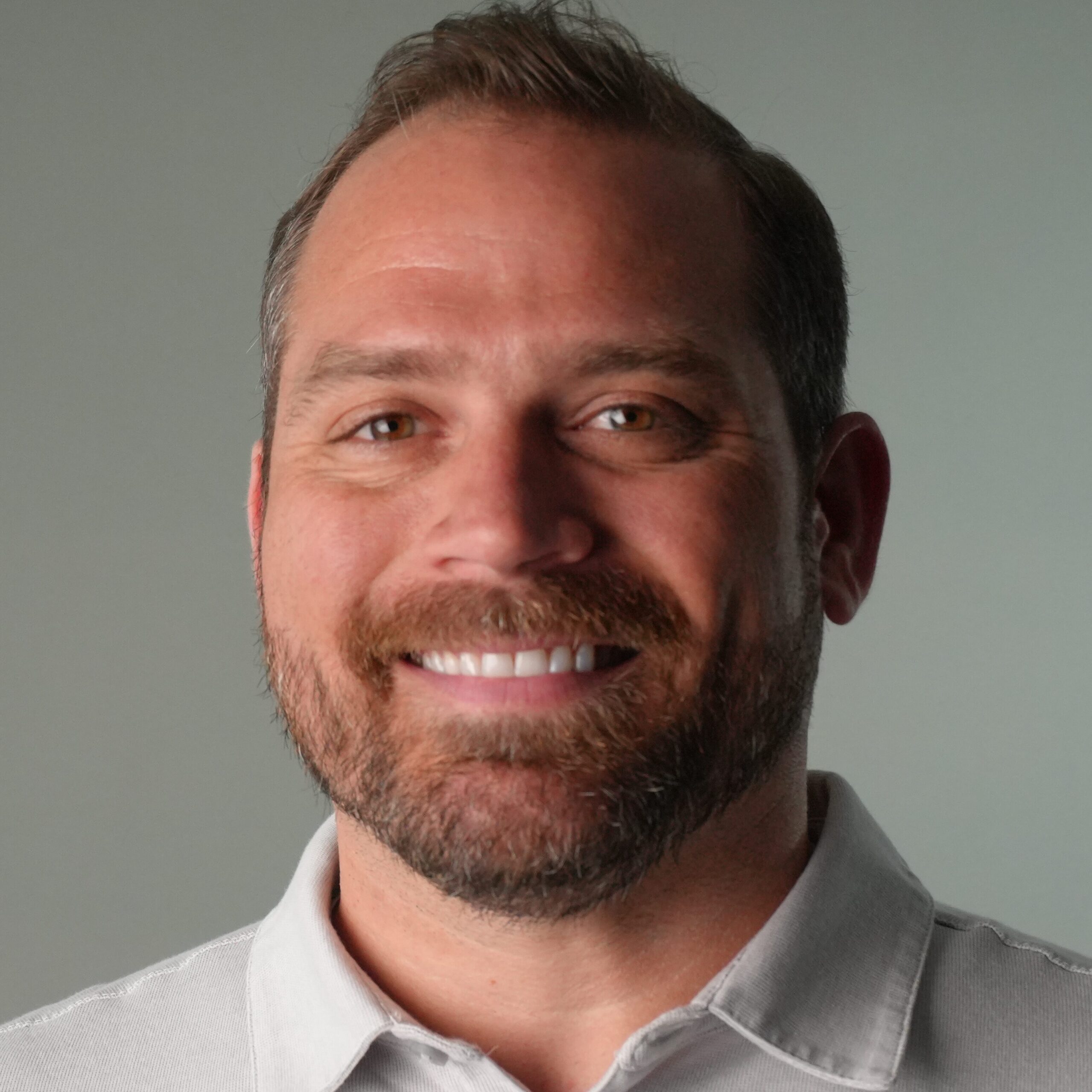Close-up of a smiling man wearing a light gray collared shirt, showing his mouth and beard.