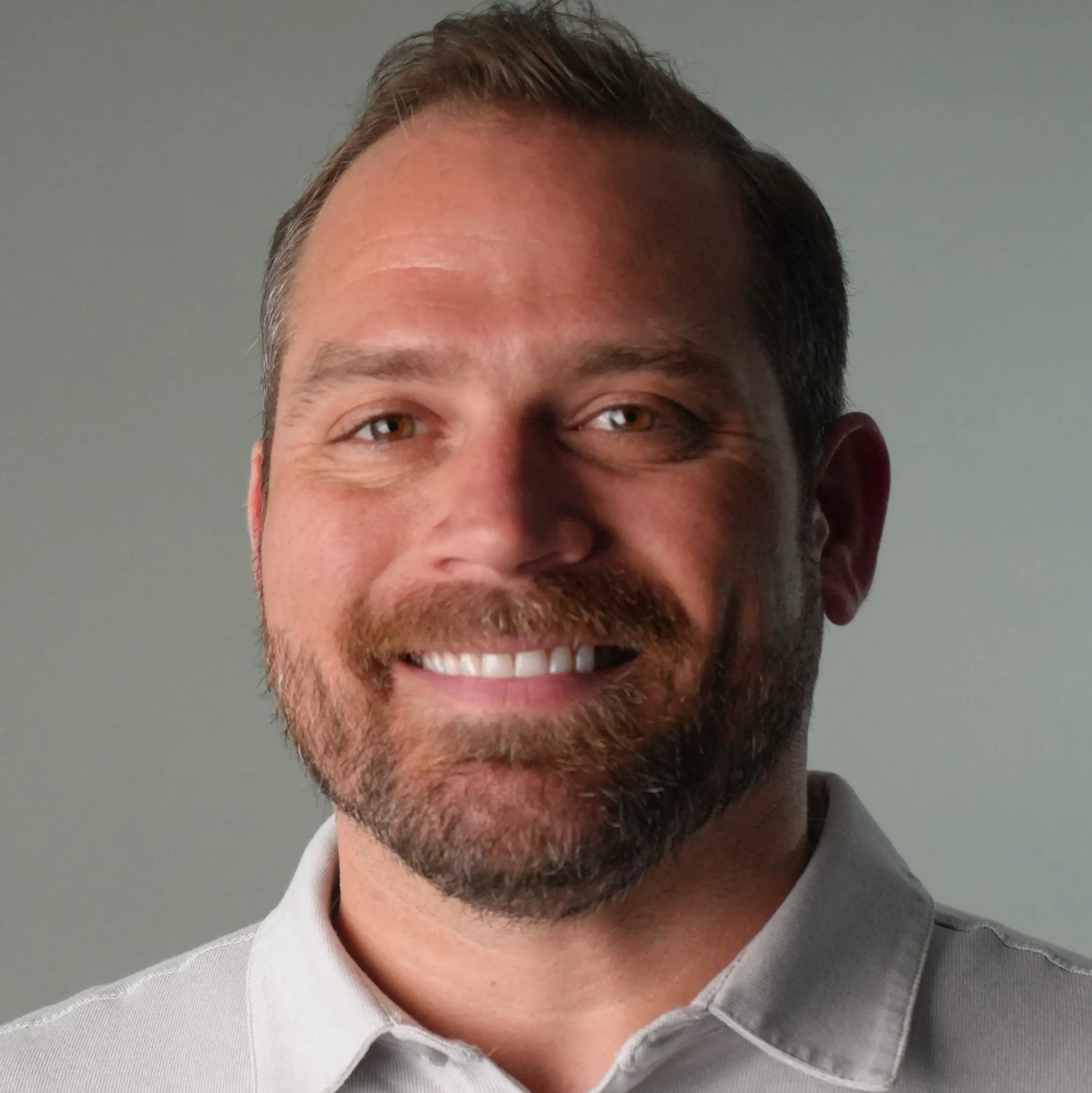 Close-up of a smiling man wearing a light gray collared shirt, showing his mouth and beard.