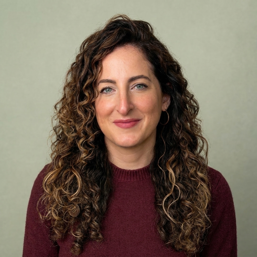Portrait of a smiling woman with long curly brown hair in a maroon sweater, against a pale green background in a head-and-shoulders shot. #informative image