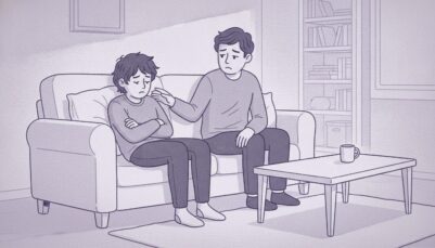 An upset teenage boy sits at home on his couch. His dad sits next to him and looks worried.